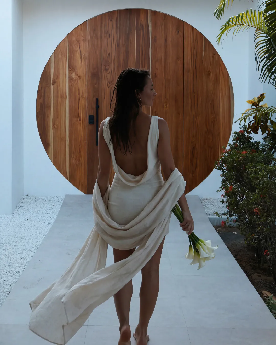 A woman in a draped linen dress walks barefoot toward a round teak door, calla lilies in hand.